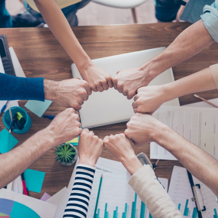 Close up cropped photo of partners putting their fists together in a circle on top of the table with work stuff. Trust, friendship, unity, cooperation concept
